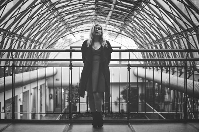 A woman in a coat stands indoors in a black and white image, striking a pose in an urban setting.