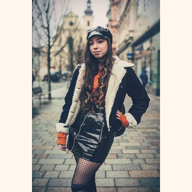 A stylish woman poses on a city street wearing a leather skirt, fishnet stockings, and jacket.