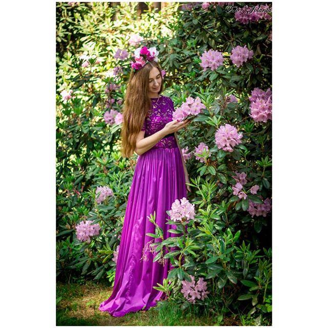 A woman in a purple dress with a flower crown stands among pink flowers in a lush garden.