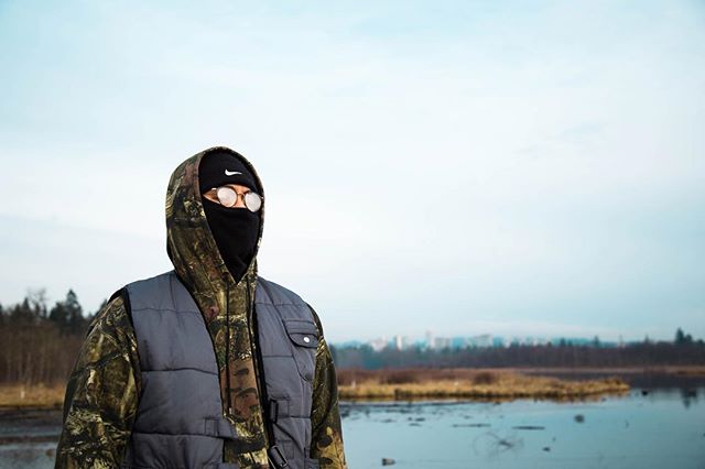 A person wearing a camouflage jacket, gray vest, and balaclava stands outdoors by a lake on a cloudy day.