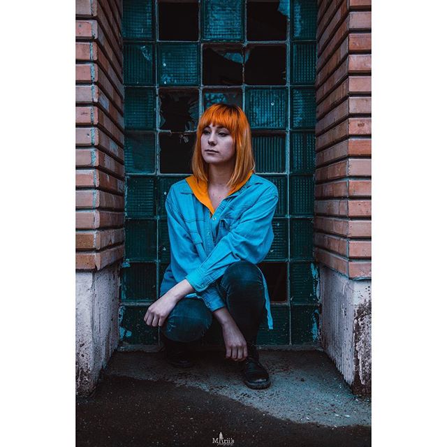 A woman with orange hair squats against a textured glass and brick wall.