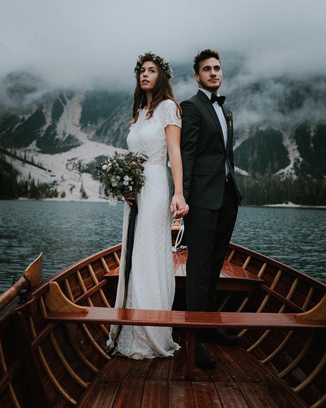 A bride and groom stand in a boat on a lake with mountains in the background.