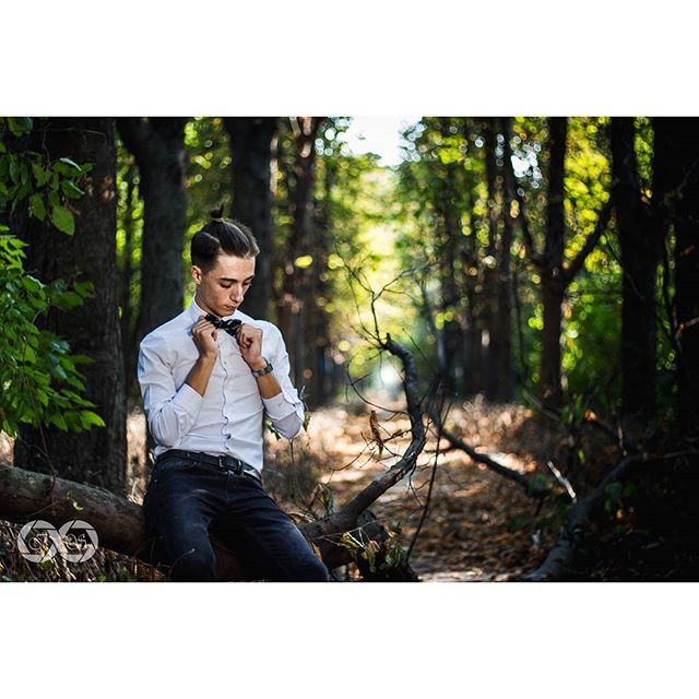 A stylish man in a bow tie sits on a log in a sun-dappled forest, embodying a blend of nature and refined fashion.