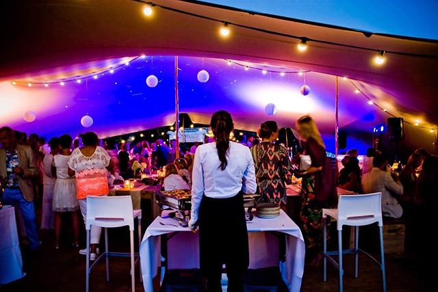A lively outdoor party reception is underway under a tent with colorful lights, tables, and people.