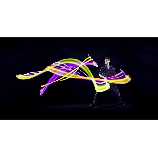 A man juggles illuminated devil sticks, creating vibrant light trails in a long exposure shot.