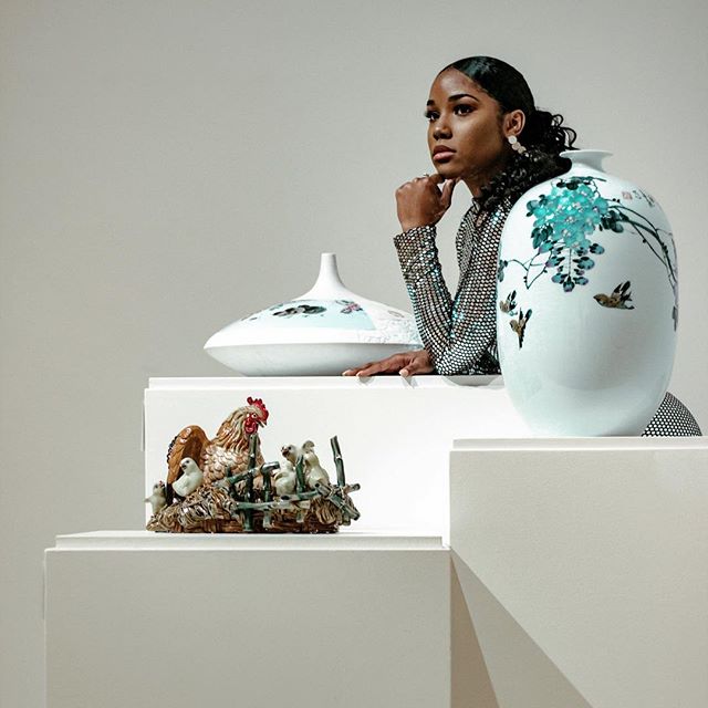 A woman poses with porcelain vases and a chicken sculpture in a studio setting, exuding elegance and artistic appreciation.