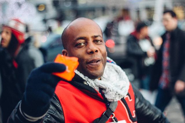 A cheerful man holds an orange object in a vibrant city street scene.