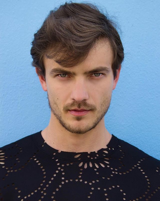 A handsome man with brown hair poses against a blue background in a black top.