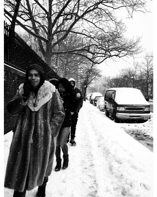 A group of people walk along a snowy sidewalk in a black and white urban setting during winter.