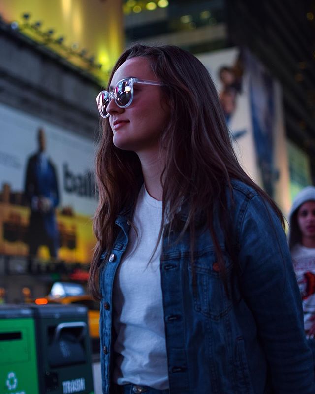 A woman in sunglasses and a denim jacket walks along a city street with billboards.