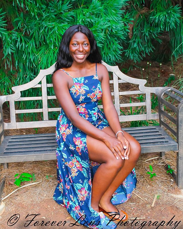 A young woman in a floral dress poses on a bench in a lush garden setting.