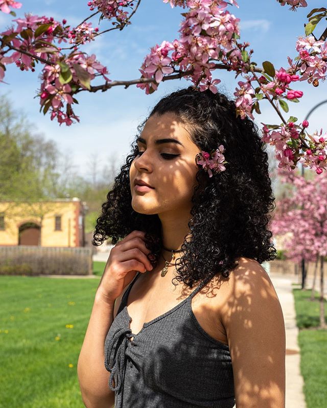 A woman with curly hair enjoys the spring weather under blooming pink blossoms in a tranquil outdoor setting.