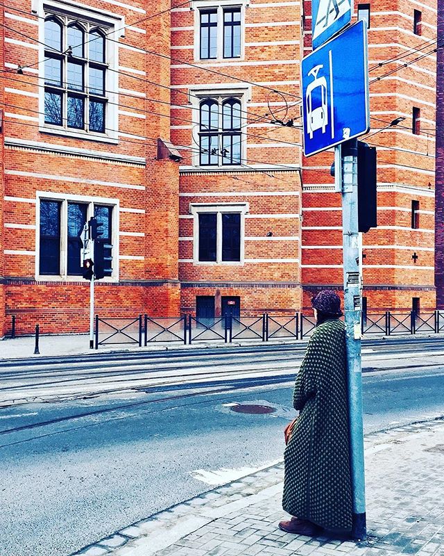 A woman stands on a city sidewalk next to a tram sign, with an old brick building in the background.