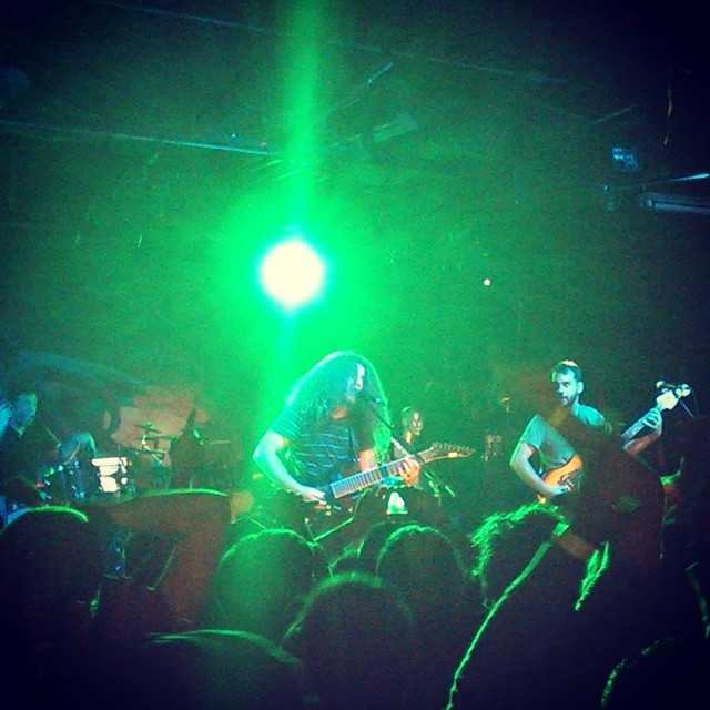 A band performs on stage under green lights, playing to an enthusiastic crowd at a live music concert.