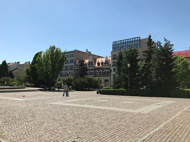 A sunny day in the city square with people walking by buildings and trees.