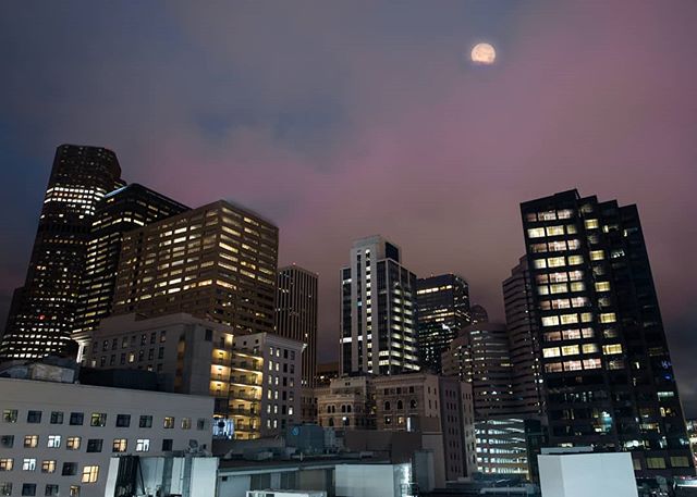A cityscape with illuminated buildings under a moody sky with a full moon shining above at night.