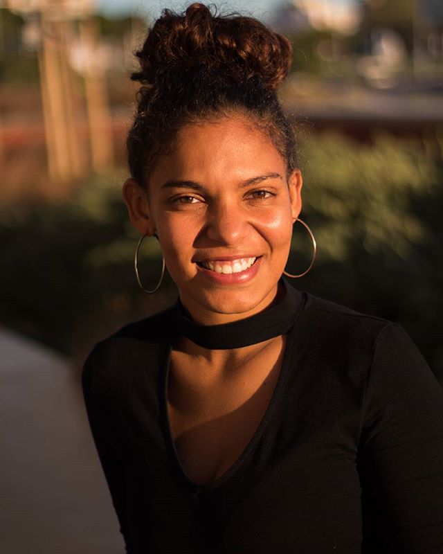 A smiling woman with a hair bun and hoop earrings poses outdoors in natural light.