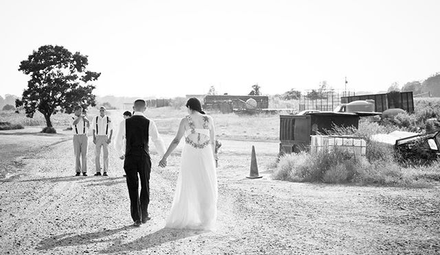 A bride and groom walk hand in hand down a country road with groomsmen in the background.