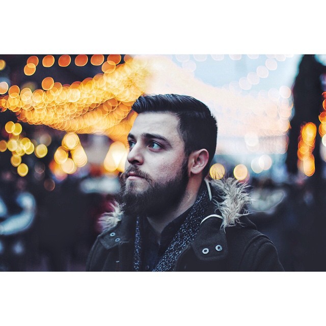 A bearded man in a coat looks off-camera with blurred city lights in the background.