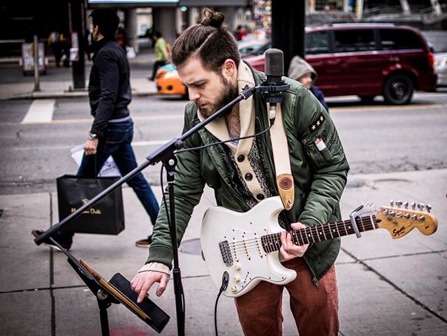 A busker plays guitar on a city street, performing for passersby with his white guitar and microphone.