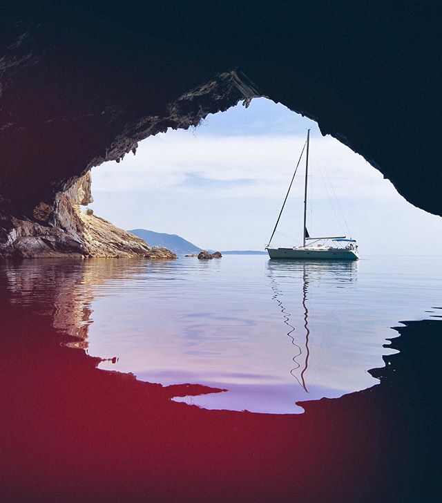 A sailboat is anchored in calm waters, framed by a scenic cave opening on a bright, cloud streaked day.