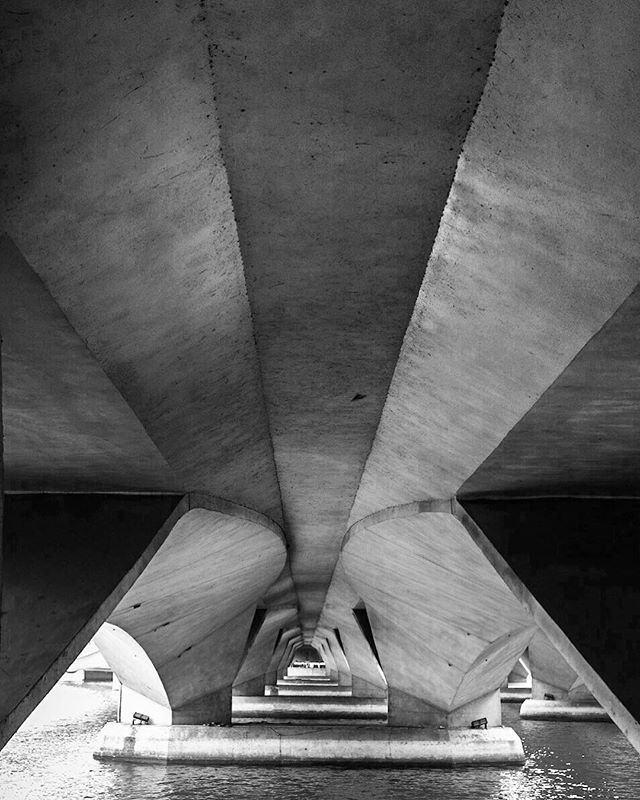 A black and white, low-angle shot reveals the geometric concrete supports of a bridge over water.