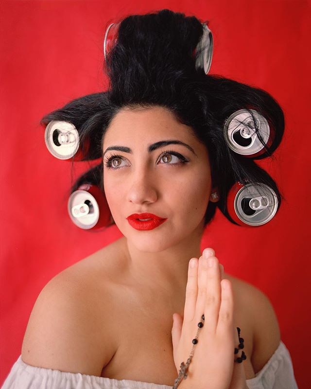 A woman with soda cans in her hair and red lipstick poses against a red background.