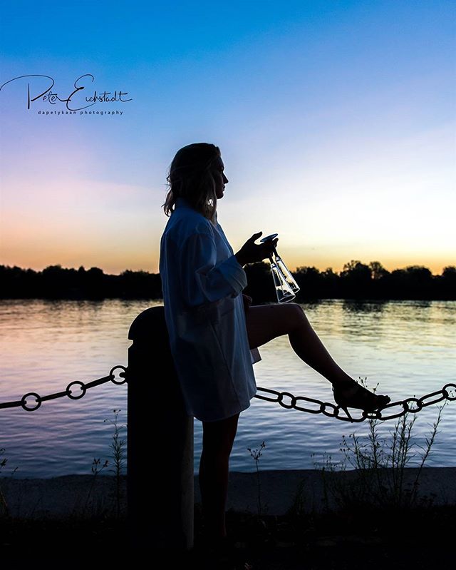 A woman is silhouetted against a colorful sunset at the river's edge, creating a tranquil scene.