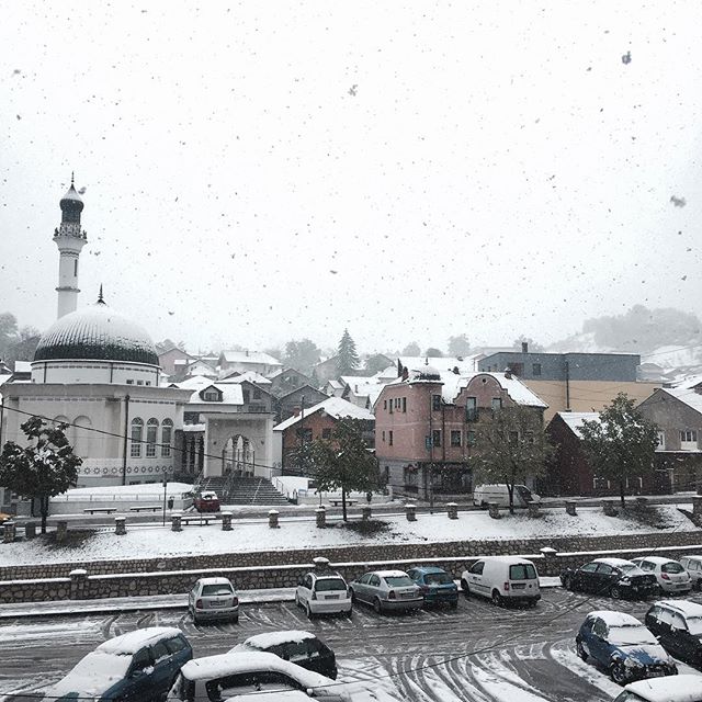 A snowy day in the city featuring a mosque, buildings and cars covered with snow.