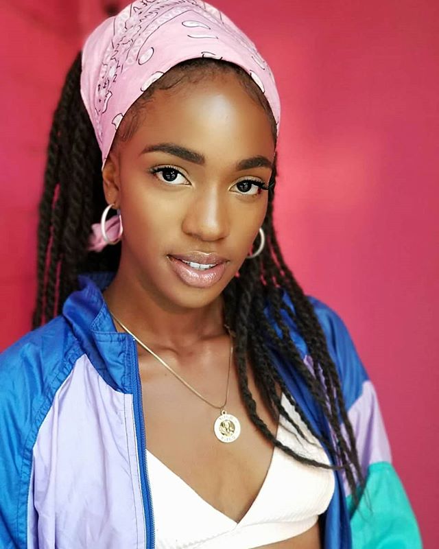 A young woman with braids and a pink bandana poses in a colorful jacket against a pink background.