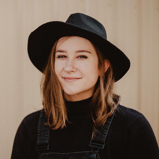 A smiling woman with a hat and overalls poses for a portrait in a neutral indoor setting.