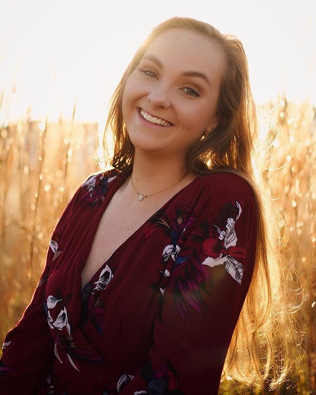 A smiling woman in a floral dress poses outdoors in a sunlit field for a portrait.