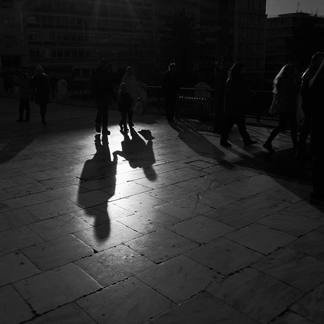 Silhouettes of people walking cast long shadows on a city street in black and white.