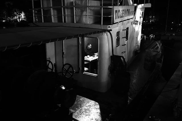 A man sits at a table on a boat at night, captured in a moody, monochrome style, evokes a sense of nostalgia and maritime history.