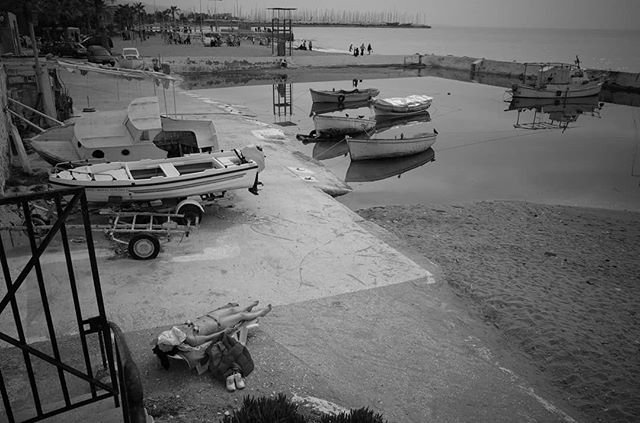 A serene black and white beach scene with boats docked and people sunbathing by the water, evokes a tranquil, vintage travel feel.