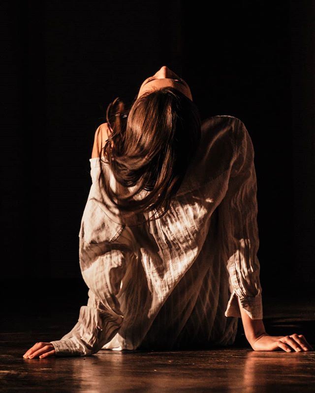 A woman in a white shirt sits looking up in a dimly lit room in an artistic, emotional pose.