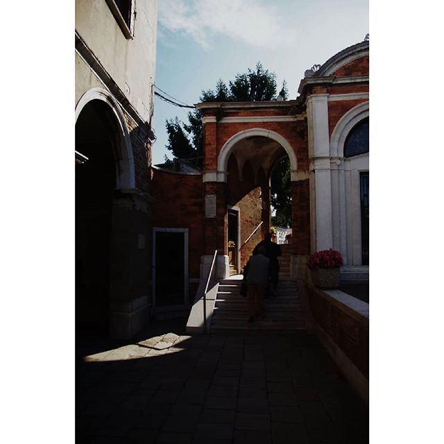People are walking up steps through an archway in an old European city, showcasing its architecture and charm.