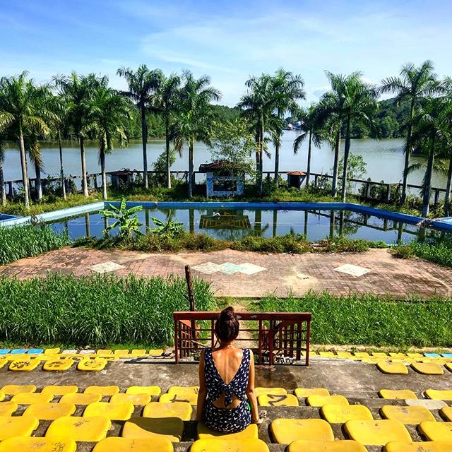 A woman sits on bleachers overlooking an abandoned pool and lake surrounded by palm trees.