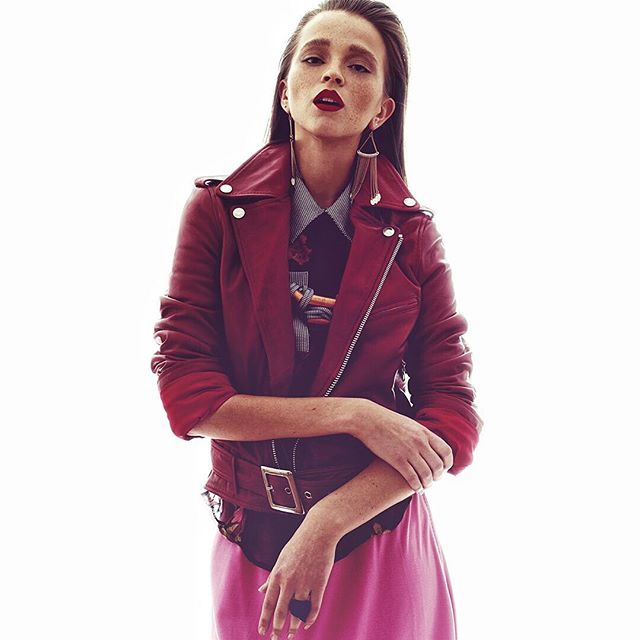 A stylish woman in a red leather jacket and pink dress poses against a white background.
