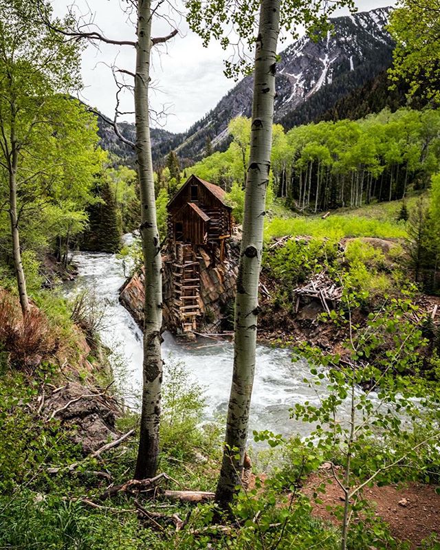 Scenic view of an old cabin beside a rushing river and waterfall nestled in lush green mountains.
