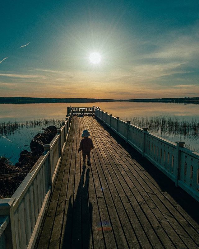 A child walks on a wooden pier toward the sun setting over a calm lake.