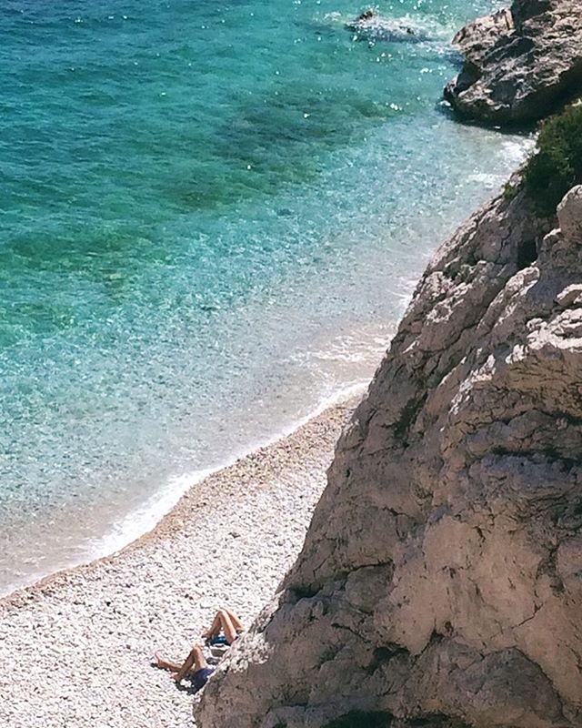 A person relaxes on a pebble beach next to clear turquoise water, framed by a rocky cliffside in a scenic coastal view.