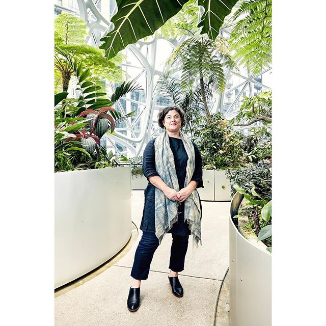 A woman stands amid lush greenery in an indoor garden setting, wearing a scarf and dark clothing.