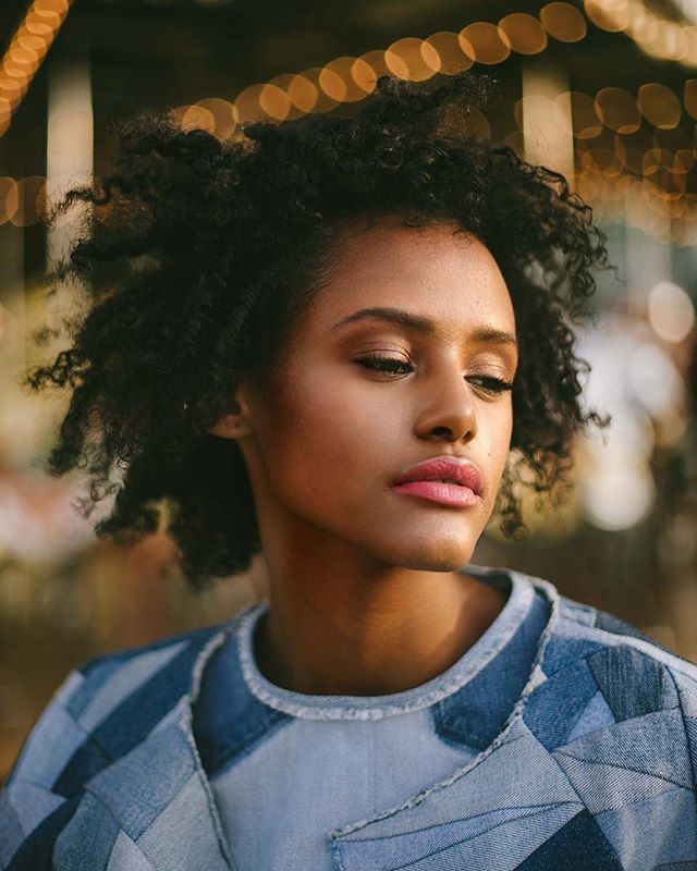 A portrait of a beautiful woman with curly hair, wearing a denim jacket, in a soft outdoor setting.