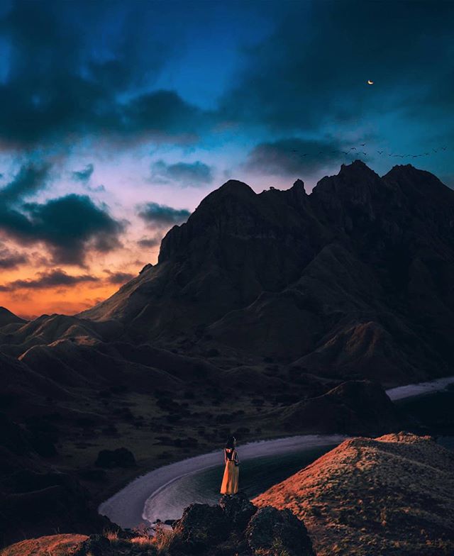 A woman stands atop a mountain, gazing at a vast landscape under a twilight sky with a crescent moon.