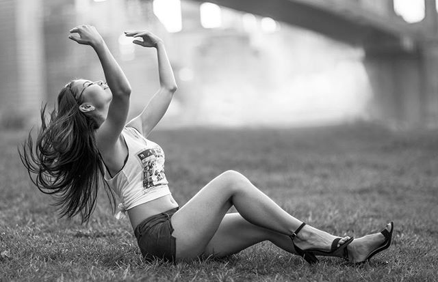 A young woman in casual attire sits on grass, striking a pose in a monochrome outdoor setting.