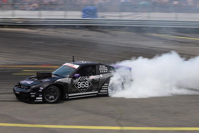 A black drift car smokes while drifting on a racetrack during a motorsport event, demonstrating power and excitement.