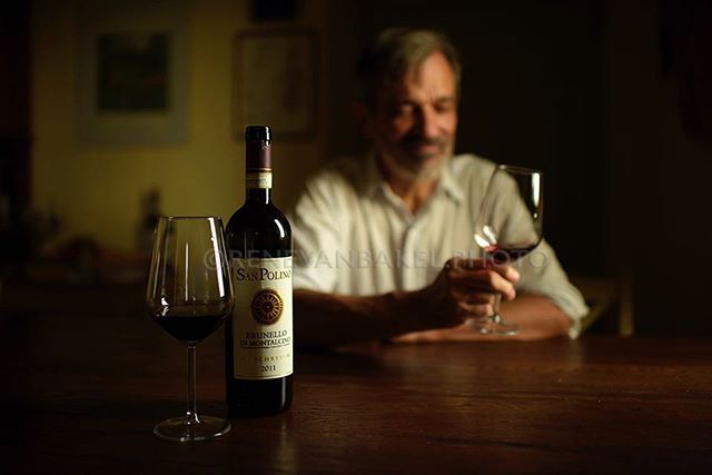 A man enjoys a glass of red wine beside a bottle of Brunello at a wooden table.