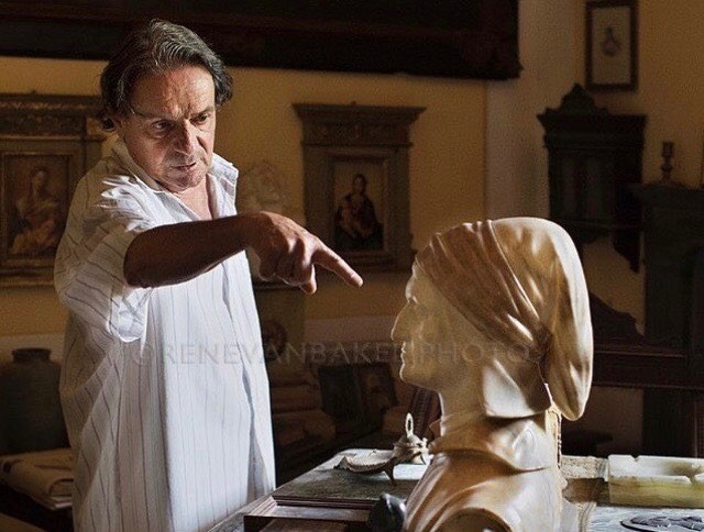 An artist stands in his studio, pointing at a bust. The soft lighting and warm tones evoke a sense of contemplation.