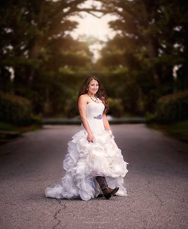 A smiling bride stands on a road wearing a wedding dress and boots for a unique country-style wedding.